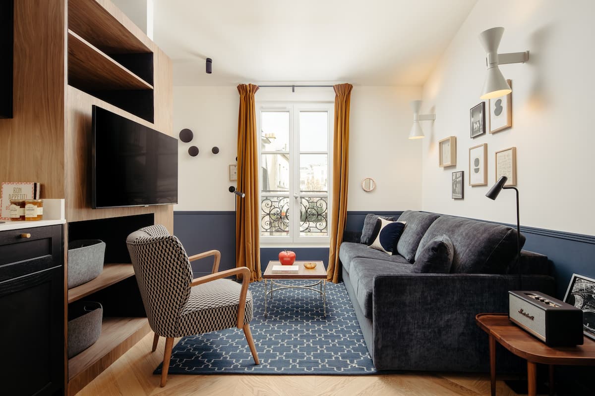 A modern living room with a flat-screen TV, a grey sofa, a patterned armchair, and a small wooden side table with a speaker.