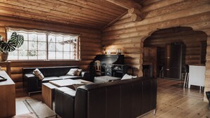 A log cabin interior with a wooden ceiling, a black leather sofa, a piano, and a large window with a view of a house outside.