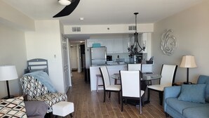 A modern living room with a sofa, a dining table with chairs, a kitchen area with cabinets and a refrigerator, and a hallway leading to another room. - Penthouse at Paradise 9th Floor (Orange Beach)