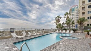 A swimming pool with lounge chairs and palm trees, surrounded by a fence and a building with balconies. - Penthouse at Paradise 9th Floor (Orange Beach)