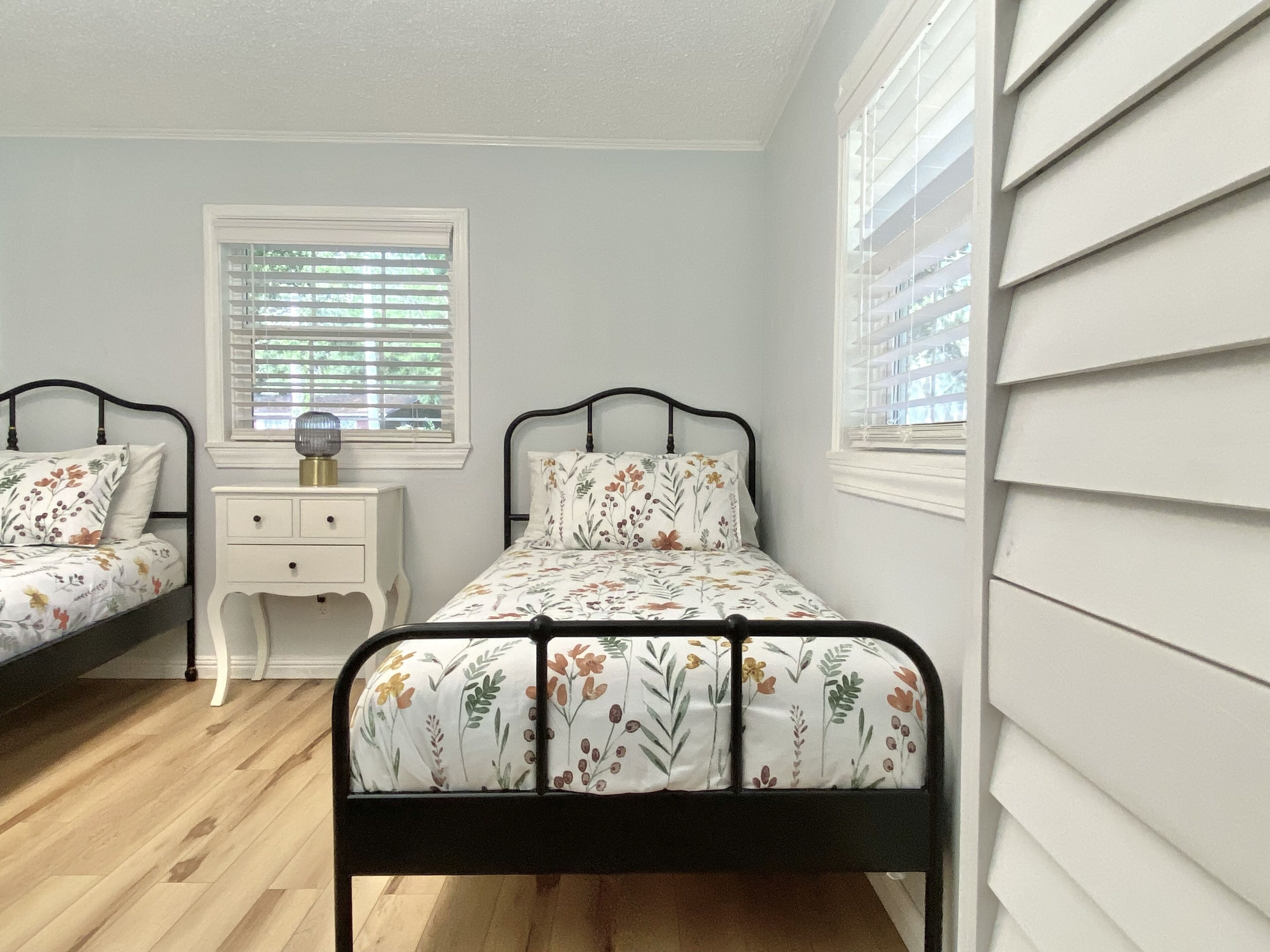 A bedroom with two single beds, a nightstand, a lamp, and a window with blinds.