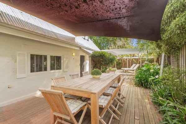 A wooden deck with a table and chairs, surrounded by greenery and a covered area.