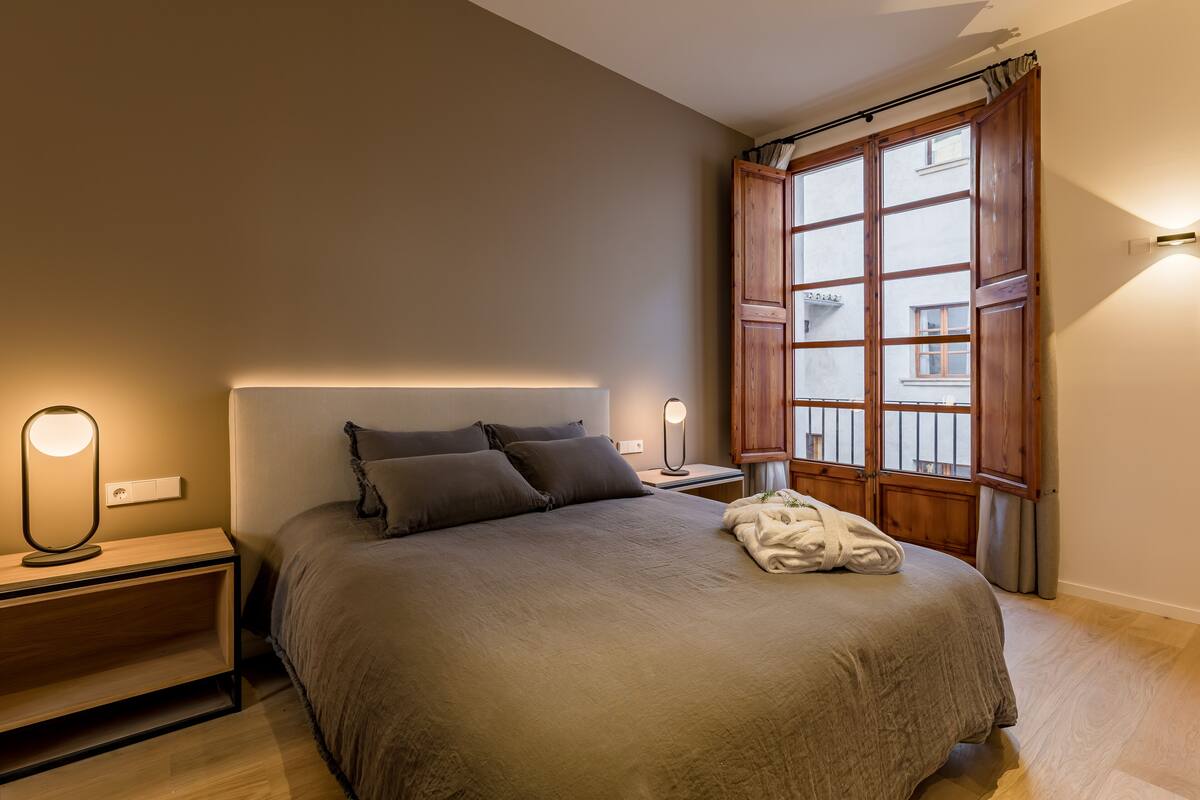 Een slaapkamer met een groot bed, twee nachtkastjes, een wandlamp en een raam met houten luiken.
