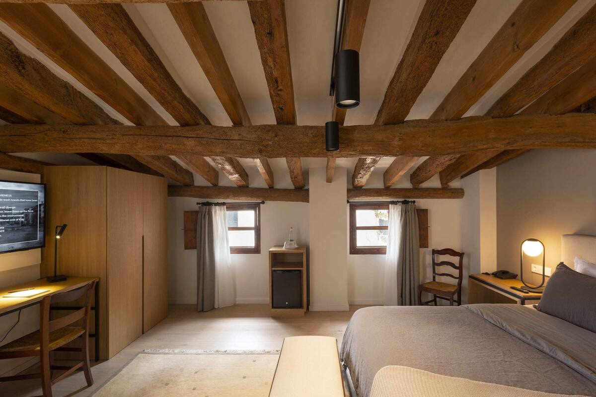 Een slaapkamer met houten plafond, een bed, een bureau, een stoel en een klein kastje.