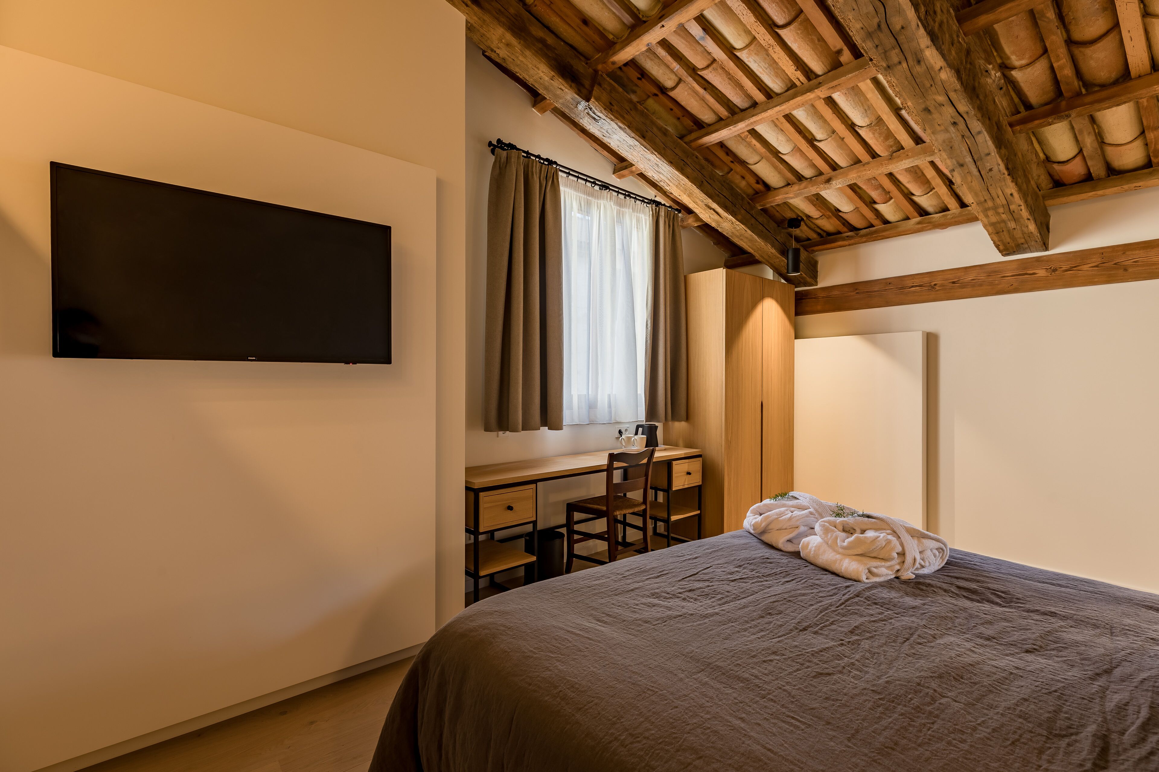 A bedroom with a bed, a desk, a chair, a TV, and a window with curtains.