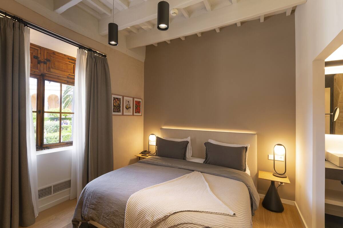 Een slaapkamer met een bed, nachtlampjes, een raam met gordijnen en een nachtkastje.