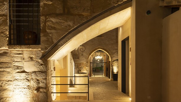 Un couloir aux murs de pierre, avec une porte en arc donnant sur un espace intérieur éclairé.