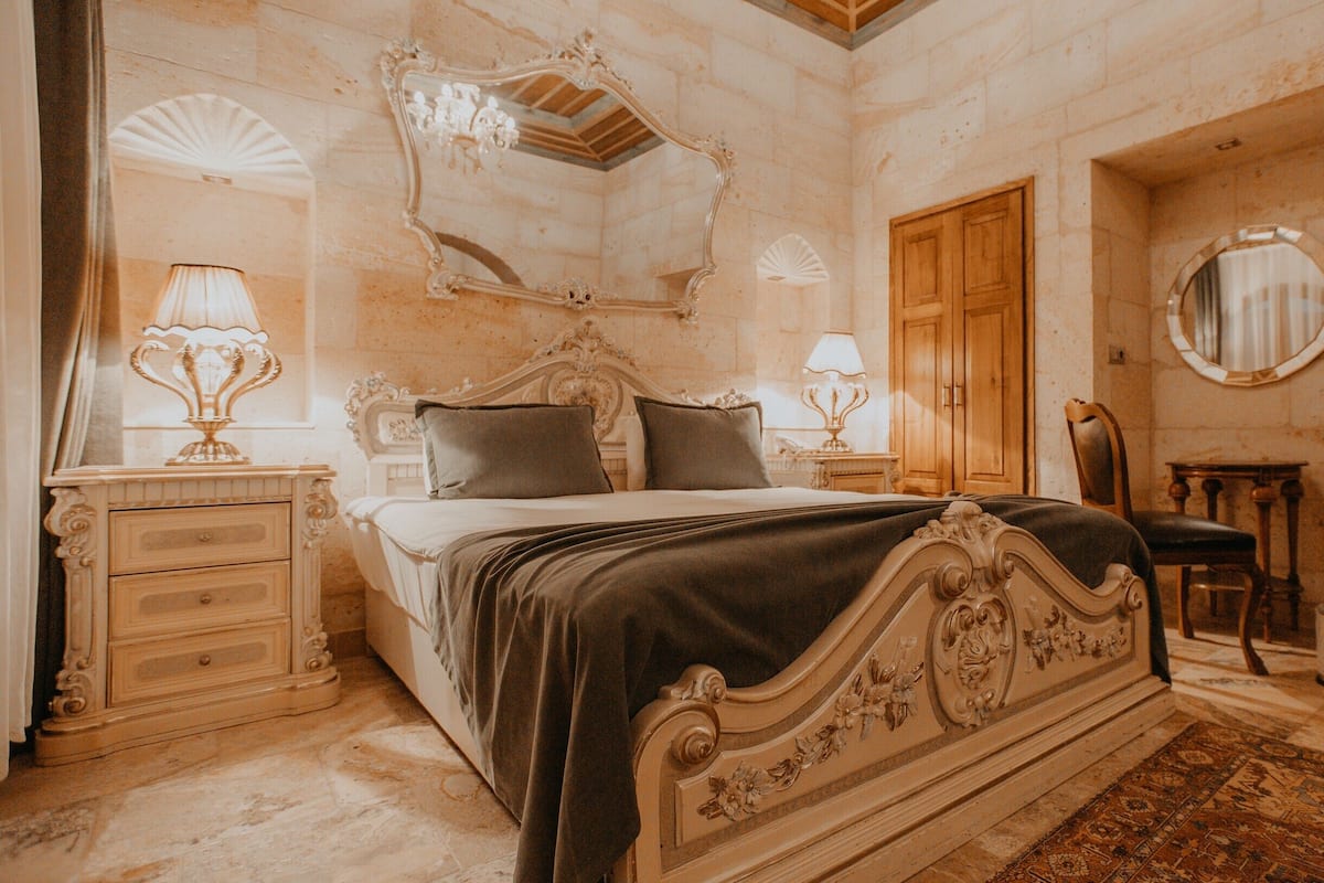 A classic bedroom with a large bed, ornate headboard, two bedside tables with lamps, a wooden wardrobe, and a mirror on the wall.