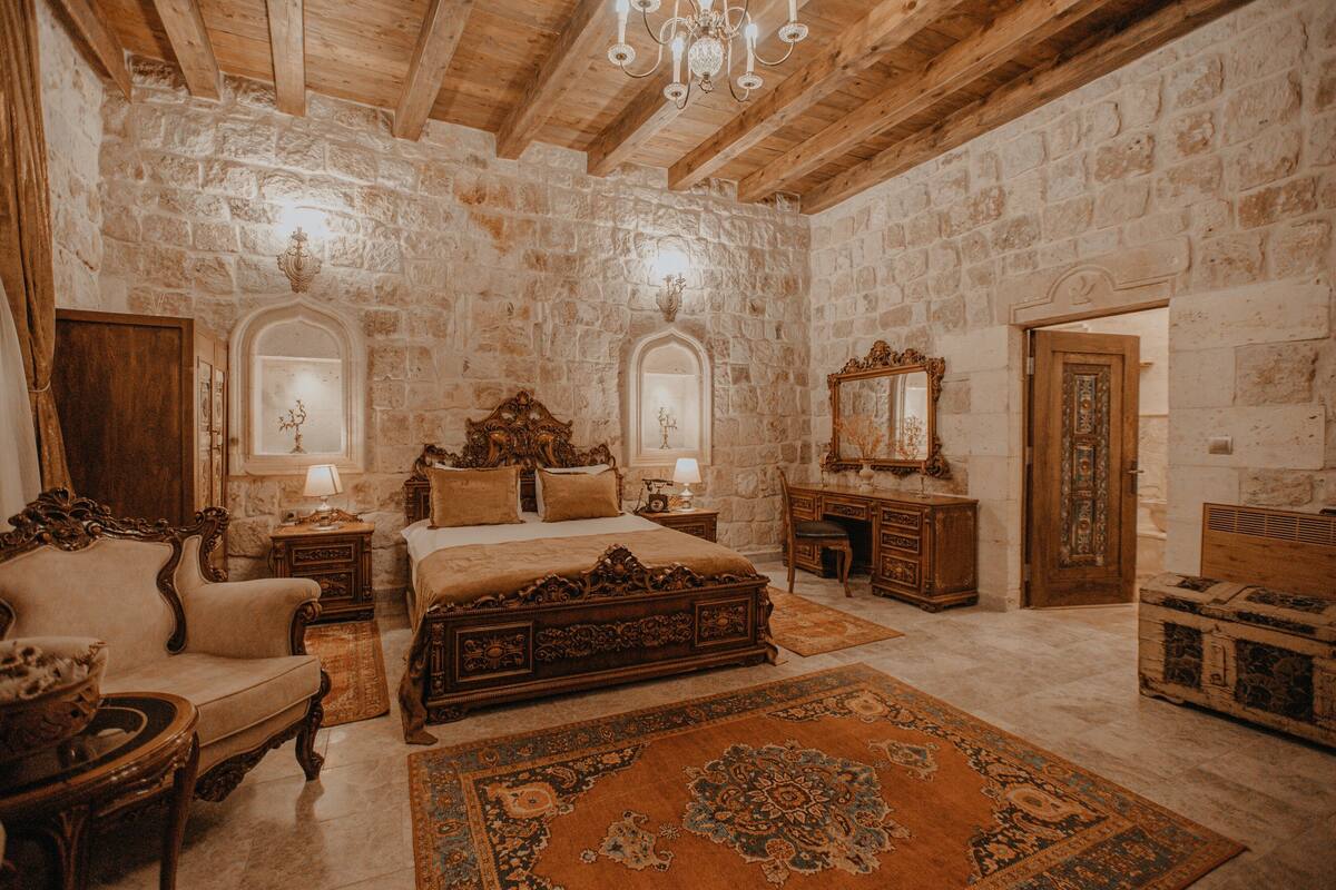 A room with a stone wall, wooden furniture, a large bed, a chandelier, and a chest of drawers.