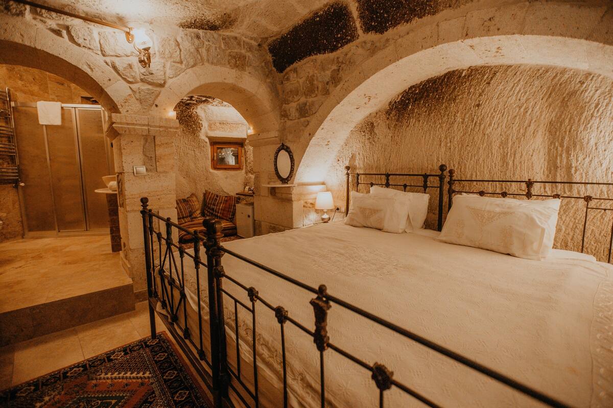 A bedroom with a large bed, a wooden headboard, a nightstand, a mirror, and a stone wall.