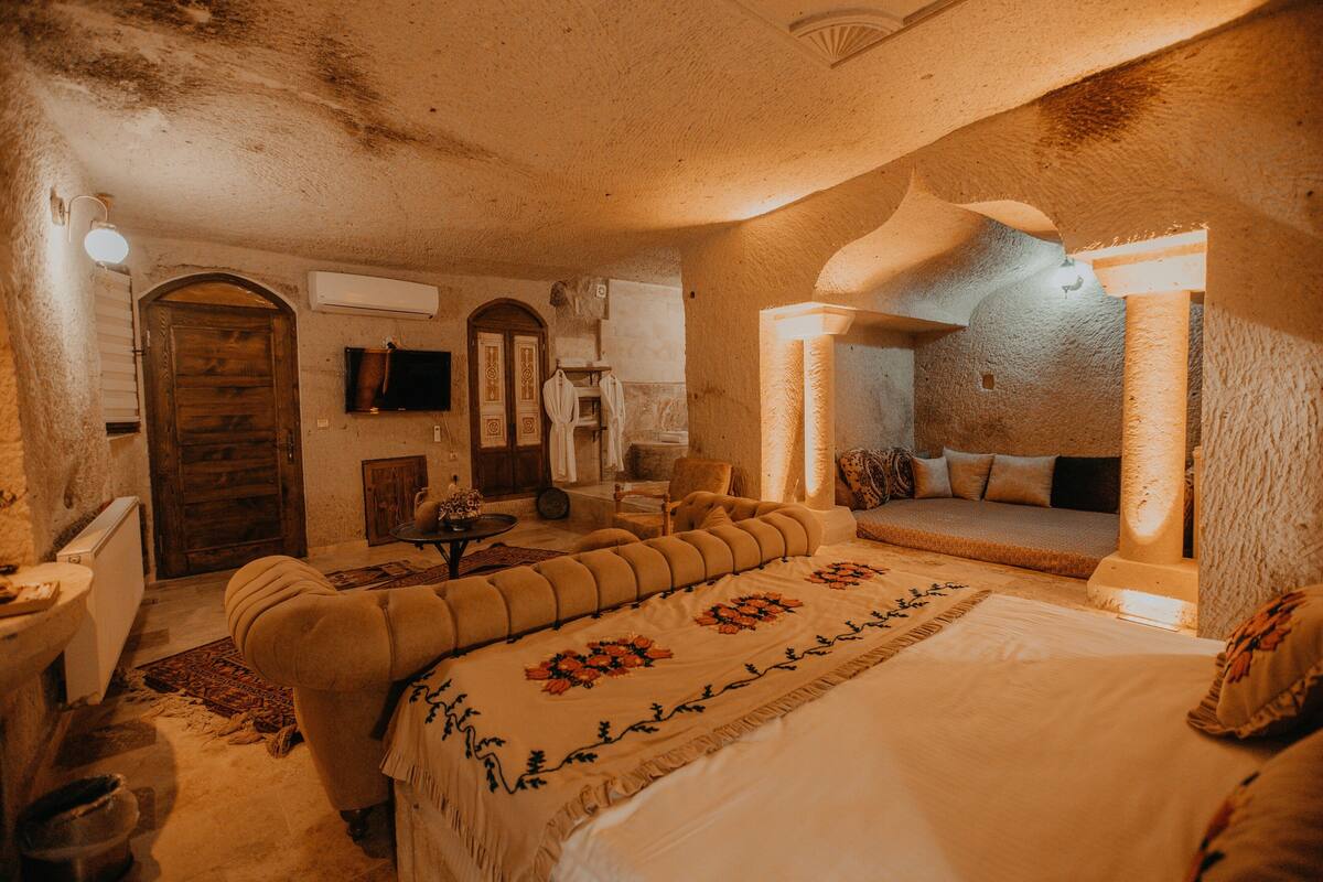 A cozy bedroom with a large bed, a sofa, and a small table. There is a wooden door and a television on the wall.