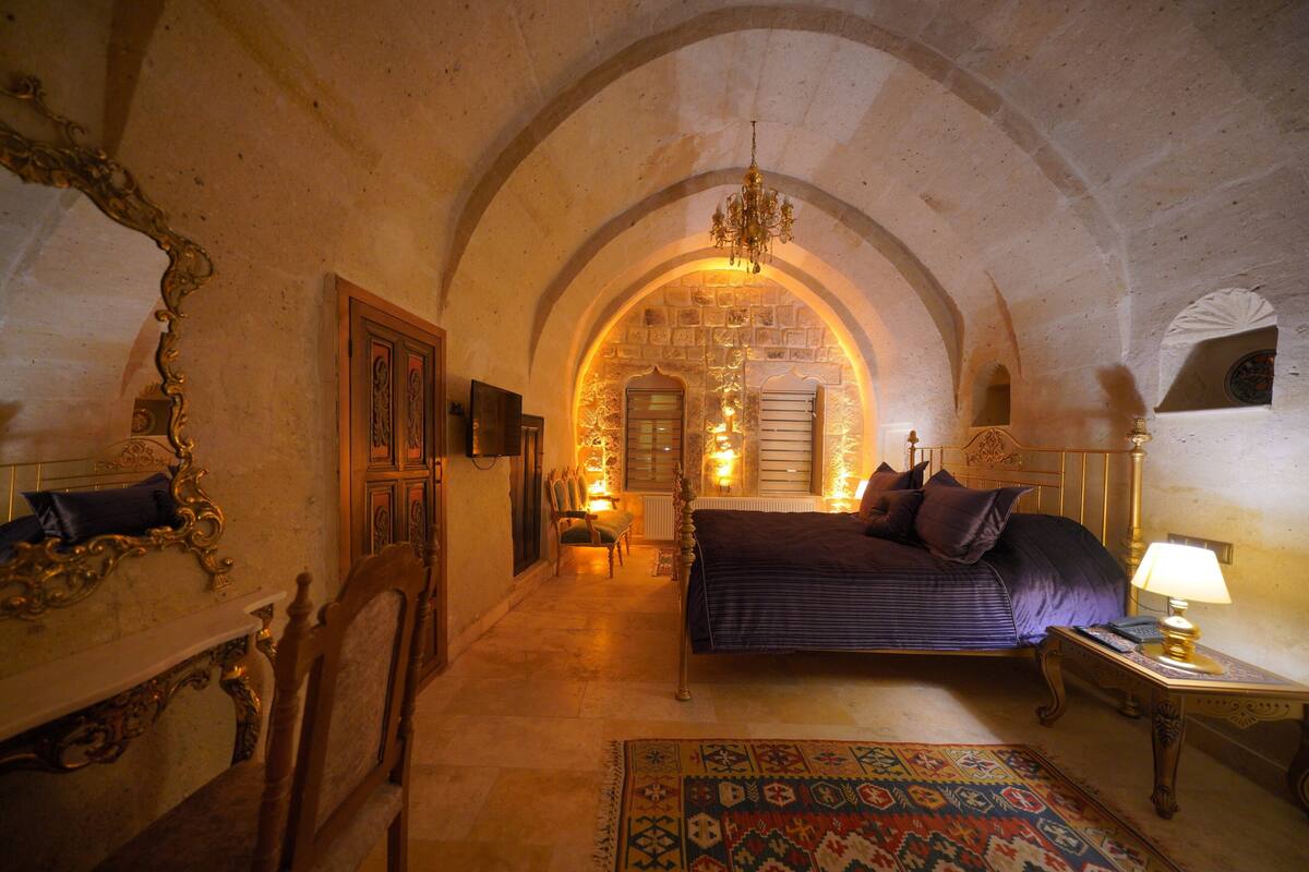 A room with an arched ceiling, a bed with a dark blue bedspread, a patterned rug, and a chandelier.