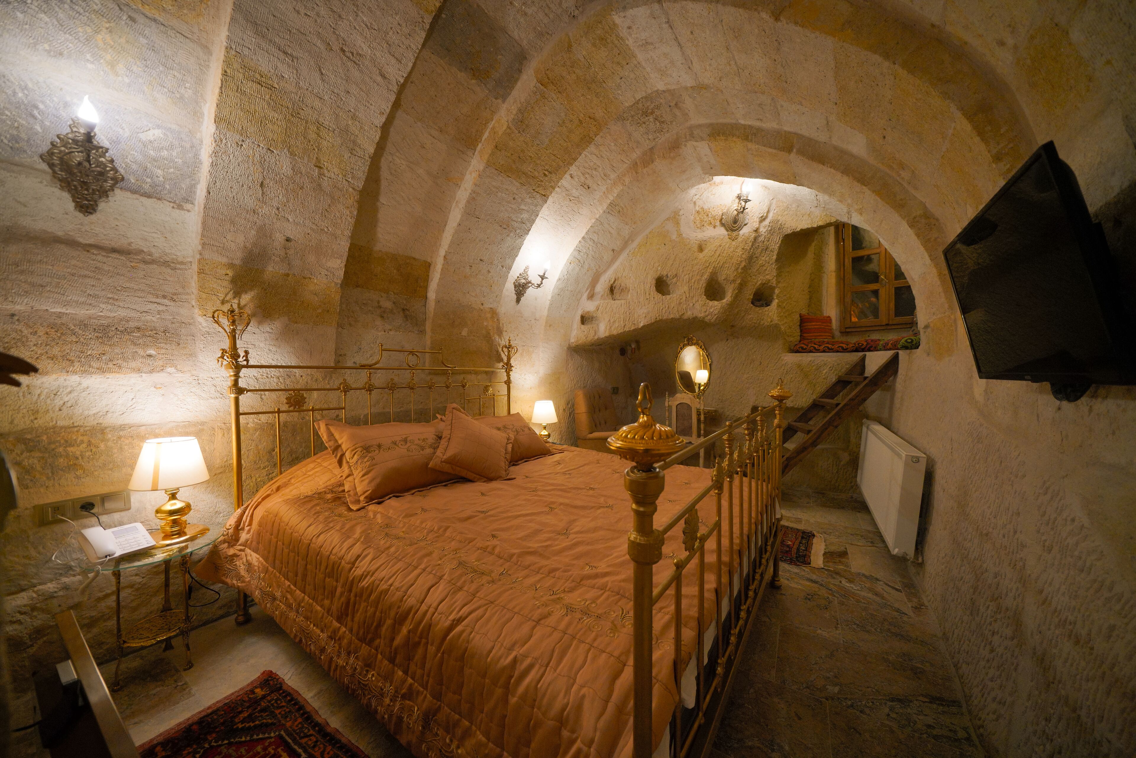 Un dormitorio con techo abovedado de piedra, una cama con cabecera, una mesita de noche con lámpara, y un televisor montado en la pared.