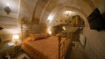 Un dormitorio con techo abovedado de piedra, una cama con cabecera, una mesita de noche con lámpara, y un televisor montado en la pared.