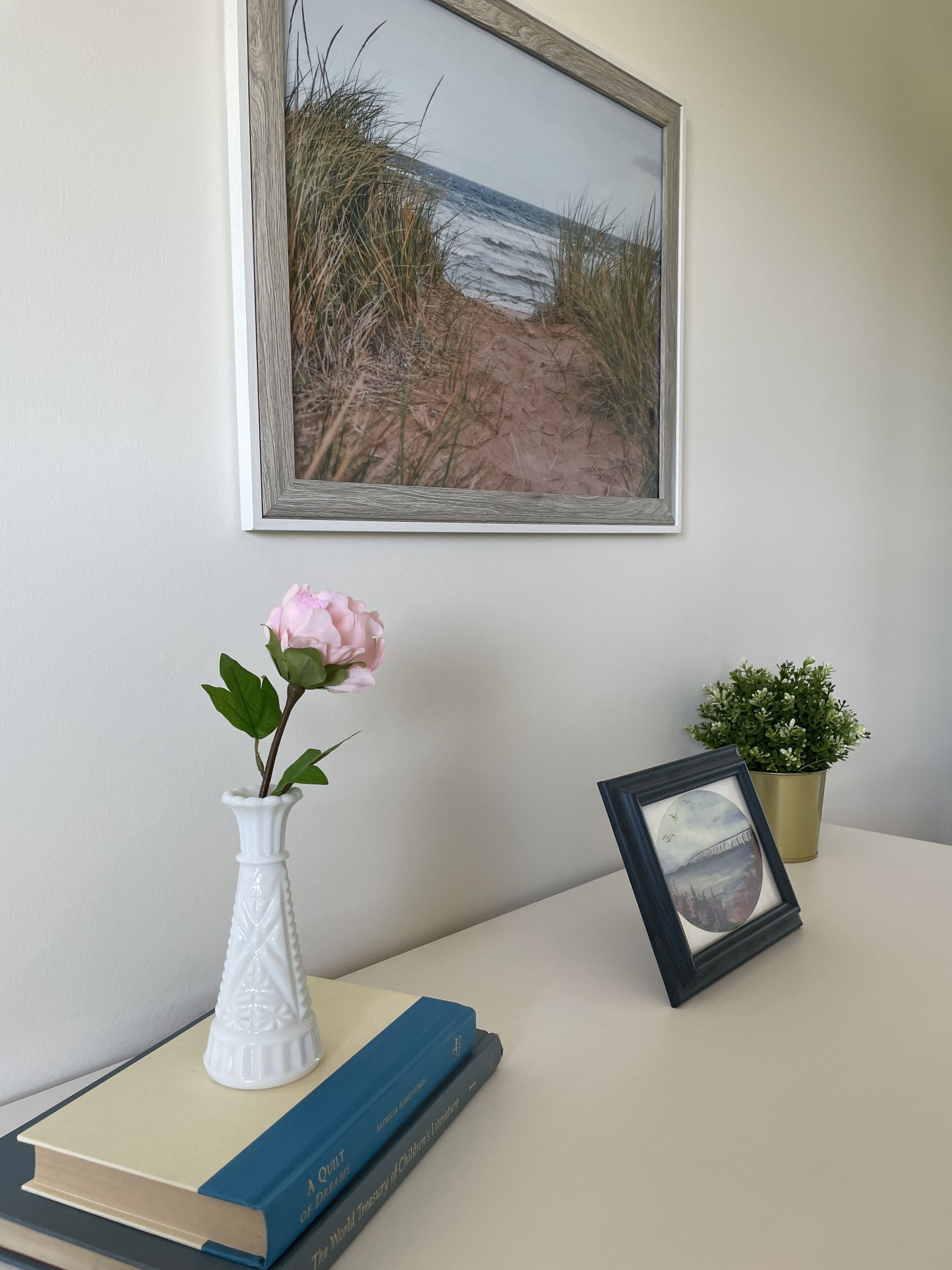 Een lijst met een schilderij van een kustlandschap, een witte vaas met een roze bloem, een kleine potplant en een lijst met een foto op een tafel.