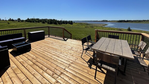 A wooden deck with outdoor furniture, including a table and chairs, overlooking a body of water and a grassy landscape. - Your Perfect Fall Getaway- Only a 5 Minute Walk From Beautiful North Shore Beach (South Rustico)