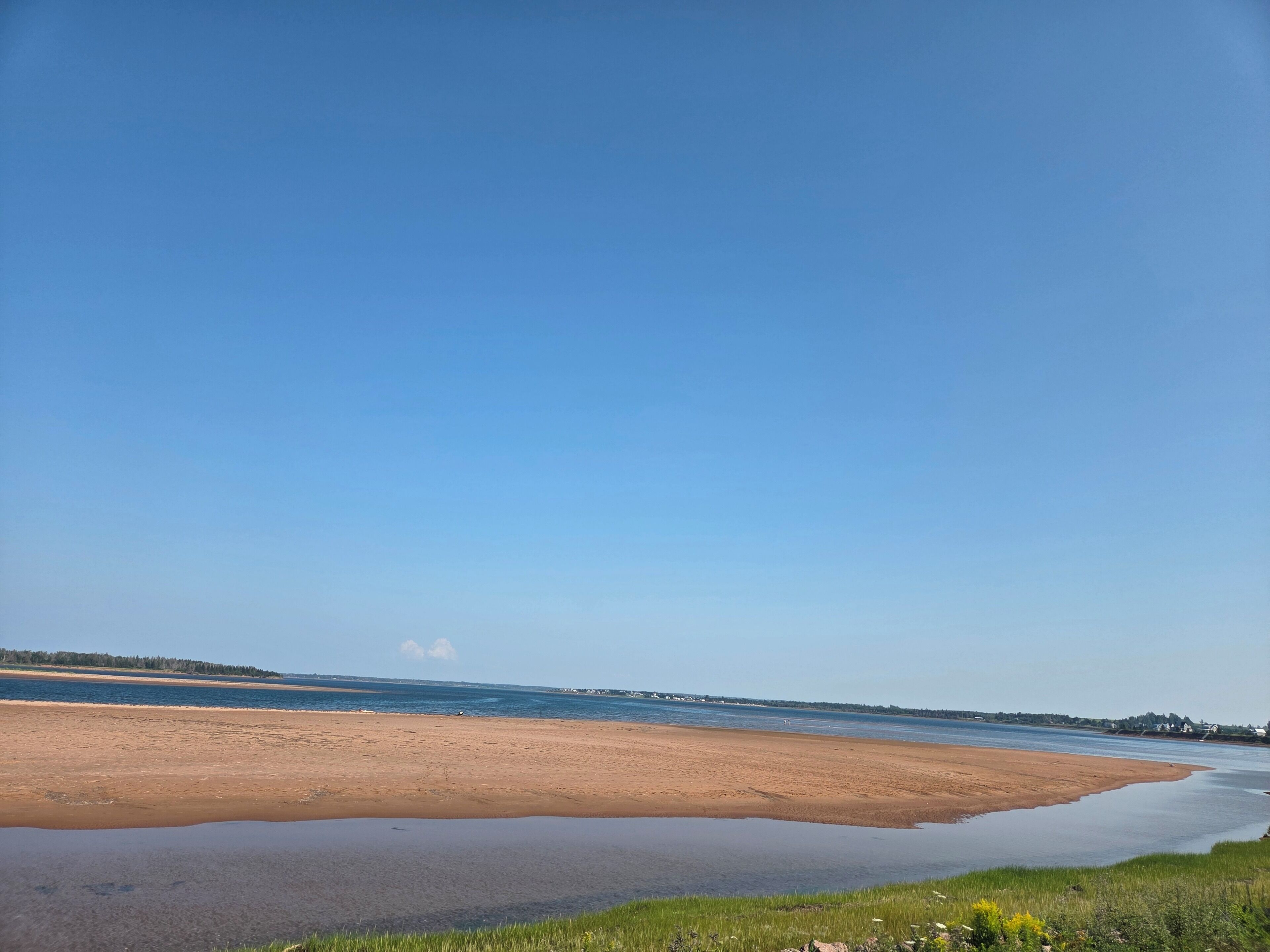 A wide sandy beach with a calm water body, a clear sky, and a distant shoreline with vegetation and buildings.
