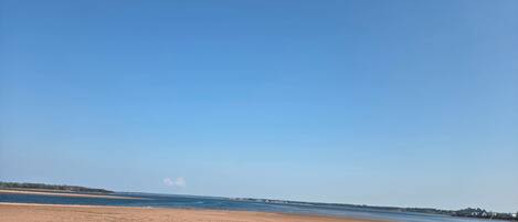 A wide sandy beach with a calm water body, a clear sky, and a distant shoreline with vegetation and buildings.