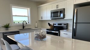 A modern kitchen with white cabinets, a stainless steel refrigerator, a microwave, and an oven. There is a large island with bar stools and a glass container of items. - Your Perfect Fall Getaway- Only a 5 Minute Walk From Beautiful North Shore Beach (South Rustico)