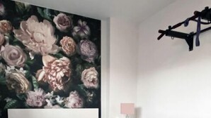 A bedroom with a bed, a floral mural, a bedside table with a lamp, and a hanging light fixture.