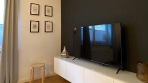 A modern living room with a flat-screen TV on a white cabinet, a small stool, and a window with curtains.