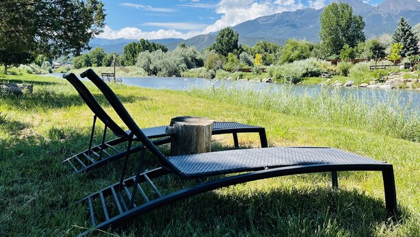 Deux fauteuils de jardin avec une table en bois, placés dans un espace verdoyant, avec vue sur un lac et des montagnes en arrière-plan.