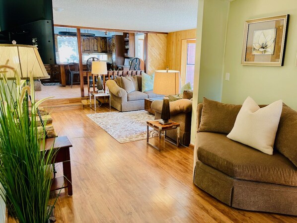 A spacious living room with a wooden floor, a brown sofa, a white armchair, and a dining area in the background.