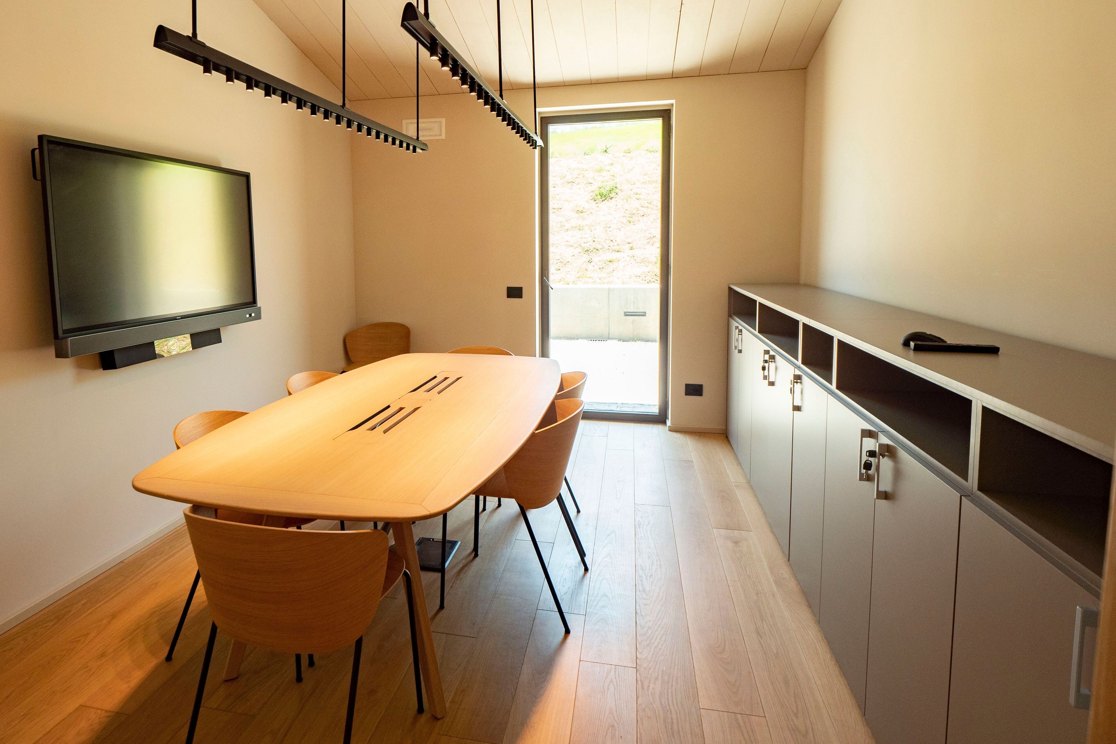 A modern kitchen with a wooden dining table, chairs, a built-in TV, and a sliding door leading to an outdoor area.