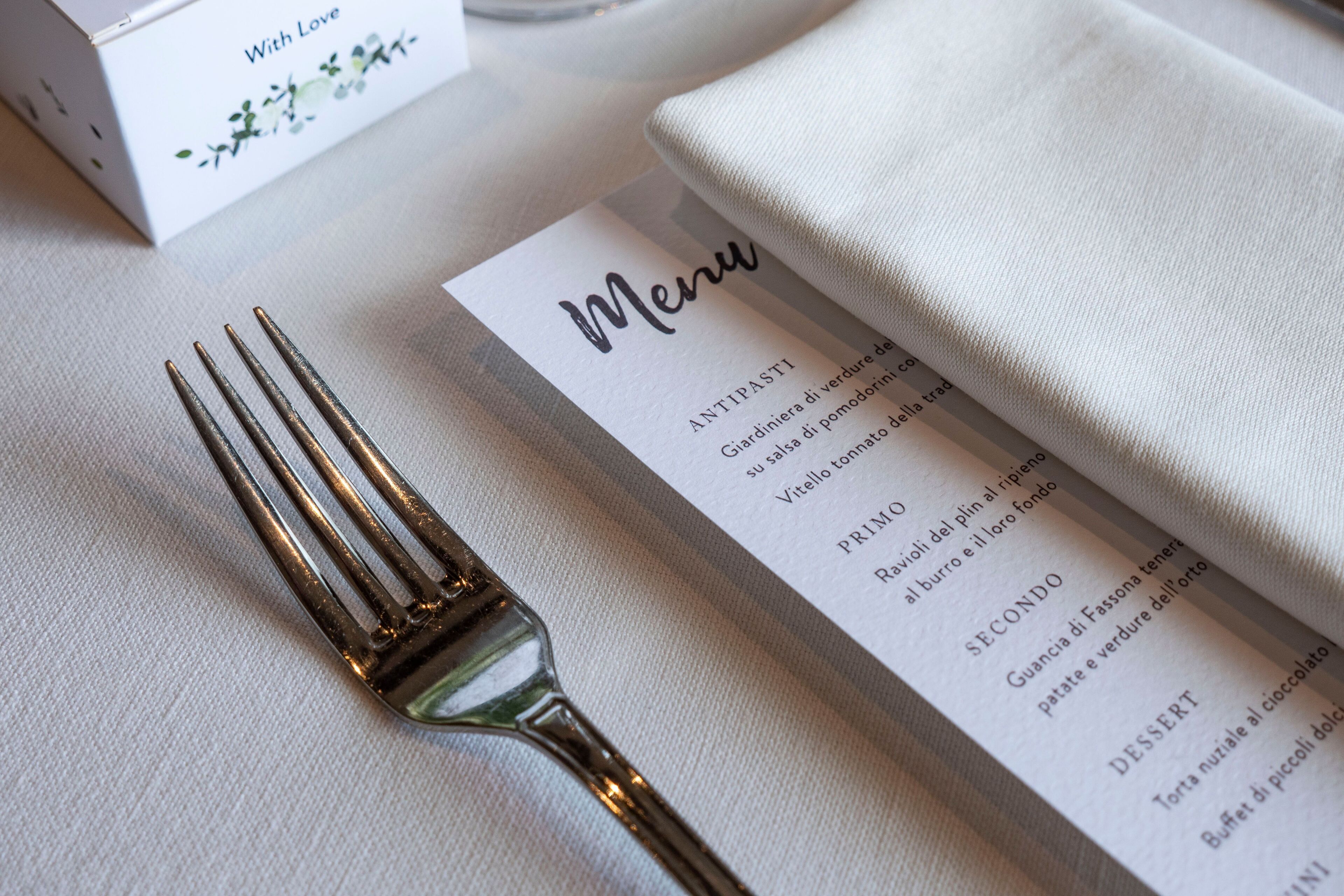 A menu with items listed in Italian, a gold-colored fork, and a white cloth napkin on a table.