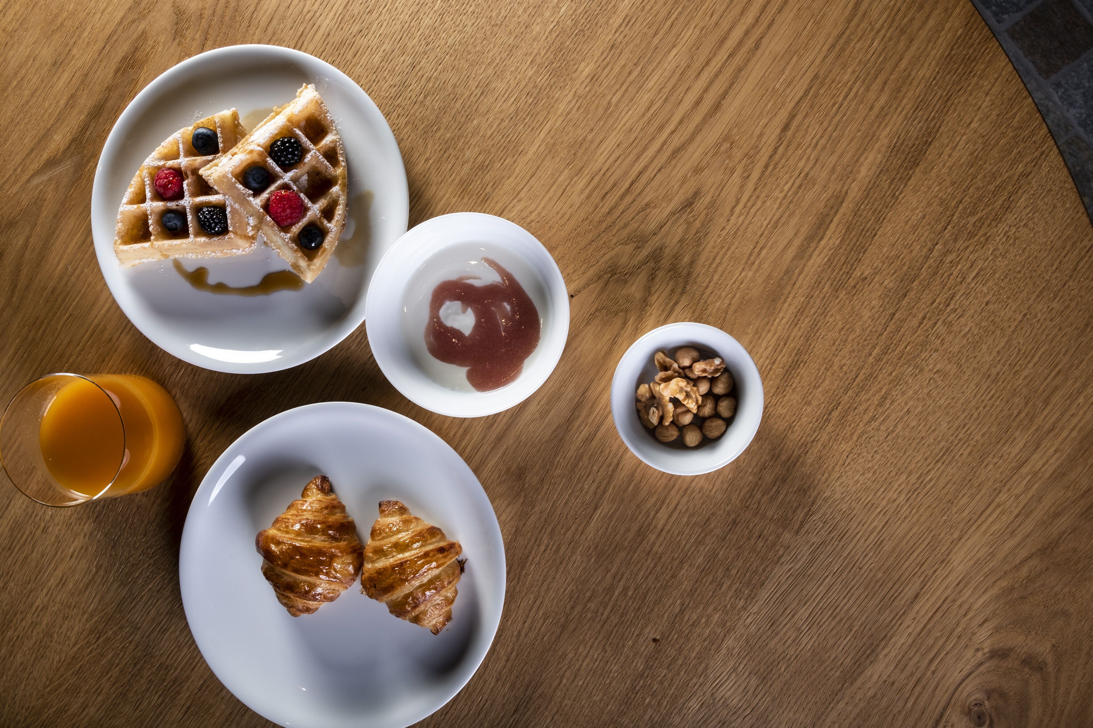 Desayuno con waffles, bayas, un croissant y un vaso de jugo de naranja sobre una mesa de madera.