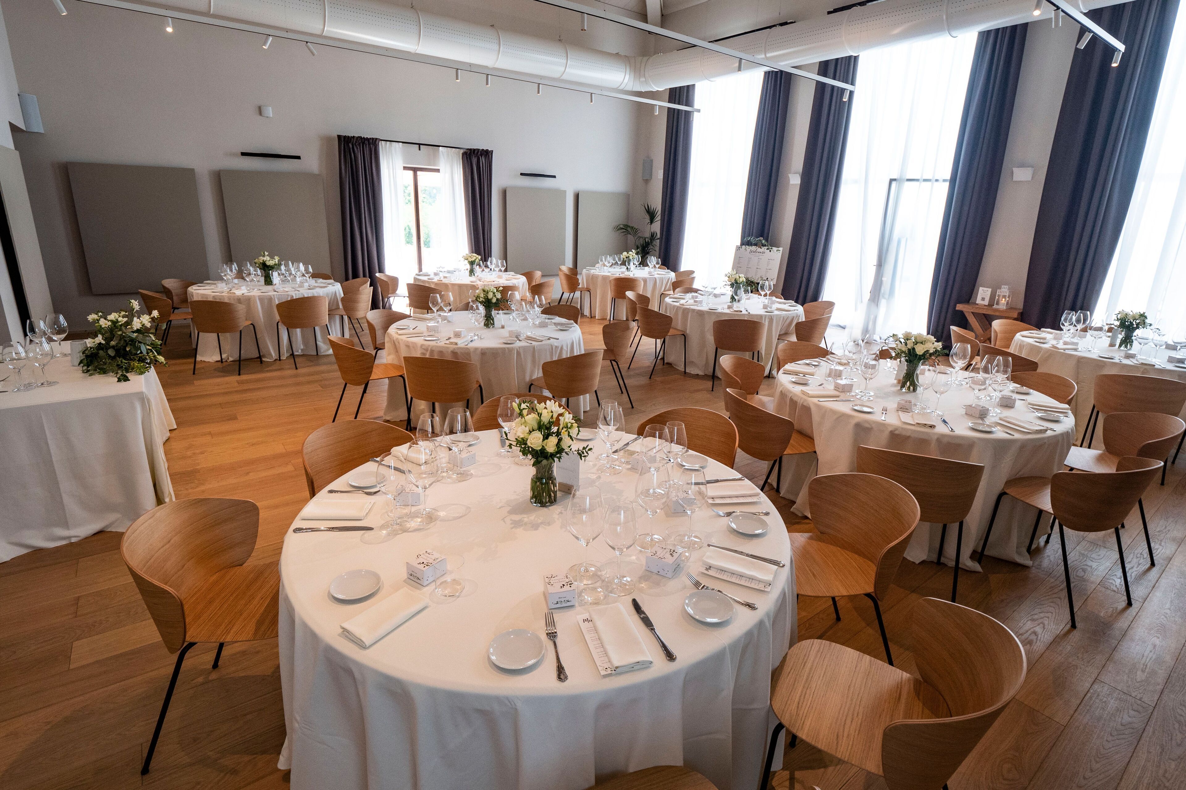 A spacious dining area with round tables set for multiple guests, featuring white tablecloths, neatly arranged cutlery, and floral centerpieces.