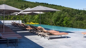 Zona de piscina con sillones de descanso y sombrillas, rodeada de un jardín ajardinado y una ladera.