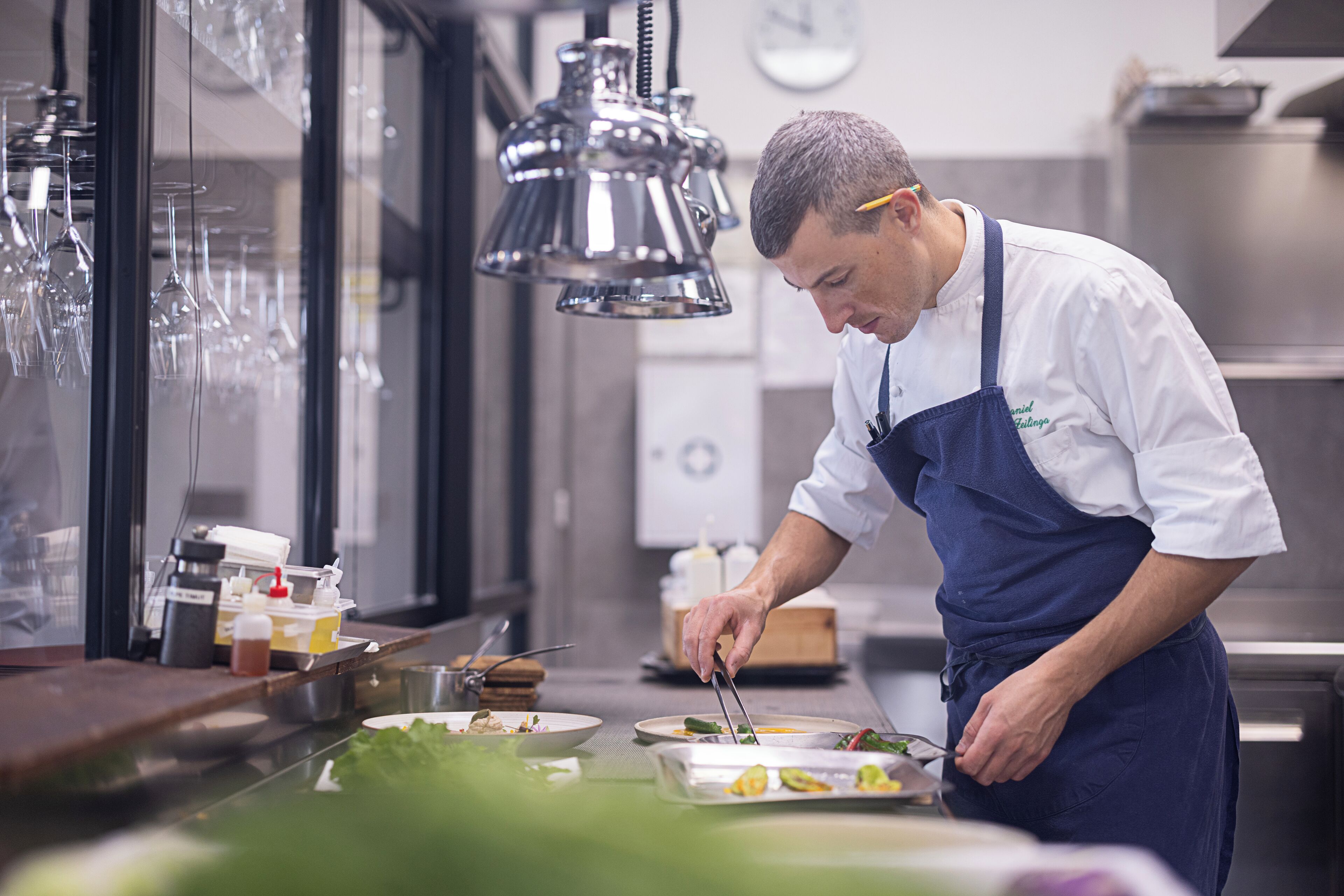 Un chef préparant des plats dans une cuisine professionnelle équipée d’appareils en acier inoxydable et où sont suspendus des verres à vin.