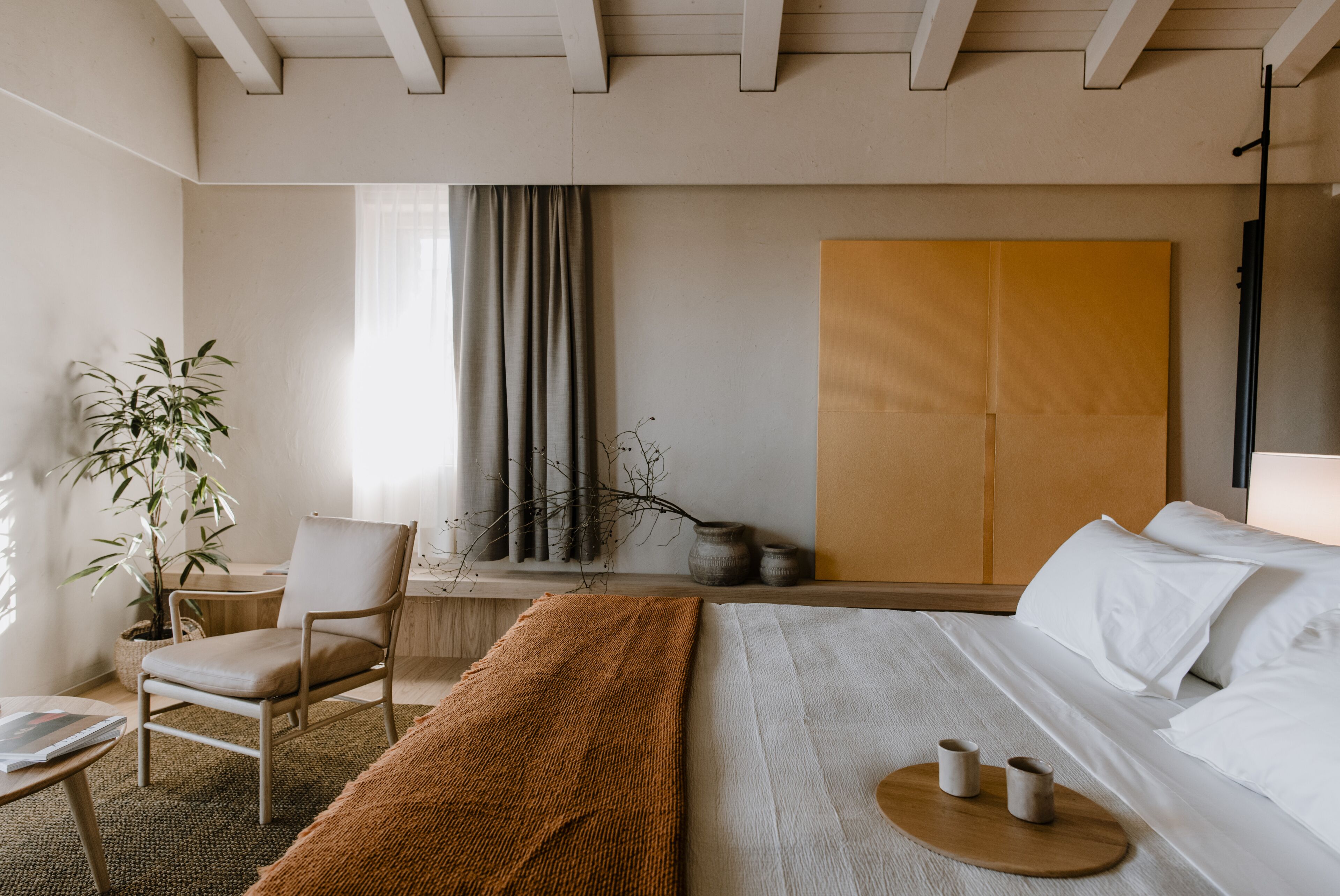 Una habitación de hotel moderna con una cama, una silla, una mesita pequeña, una planta y un ventanal grande con cortinas.