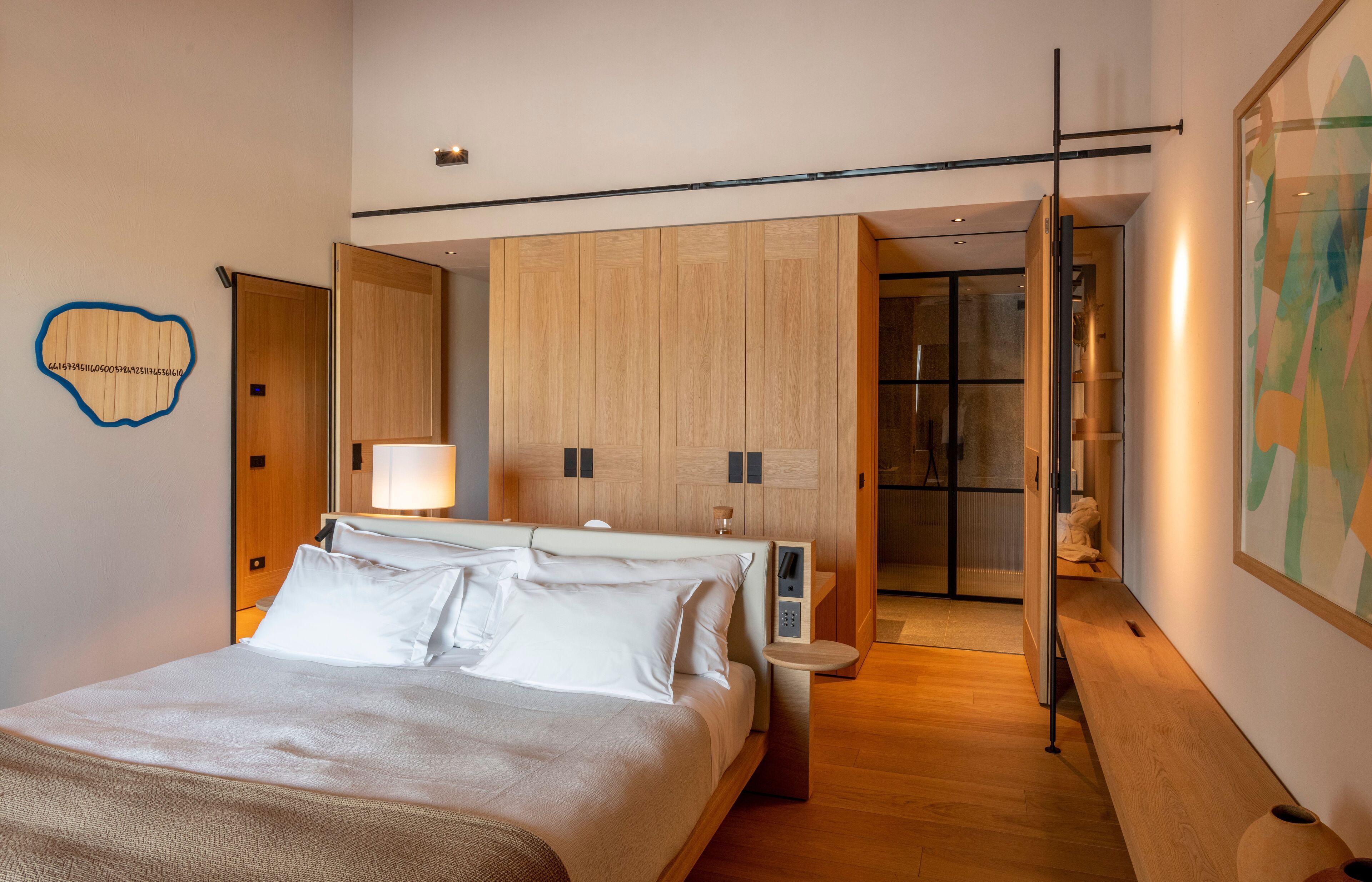 A modern bedroom with a large bed, wooden wardrobe, bedside tables, and a bathroom visible through an open door.