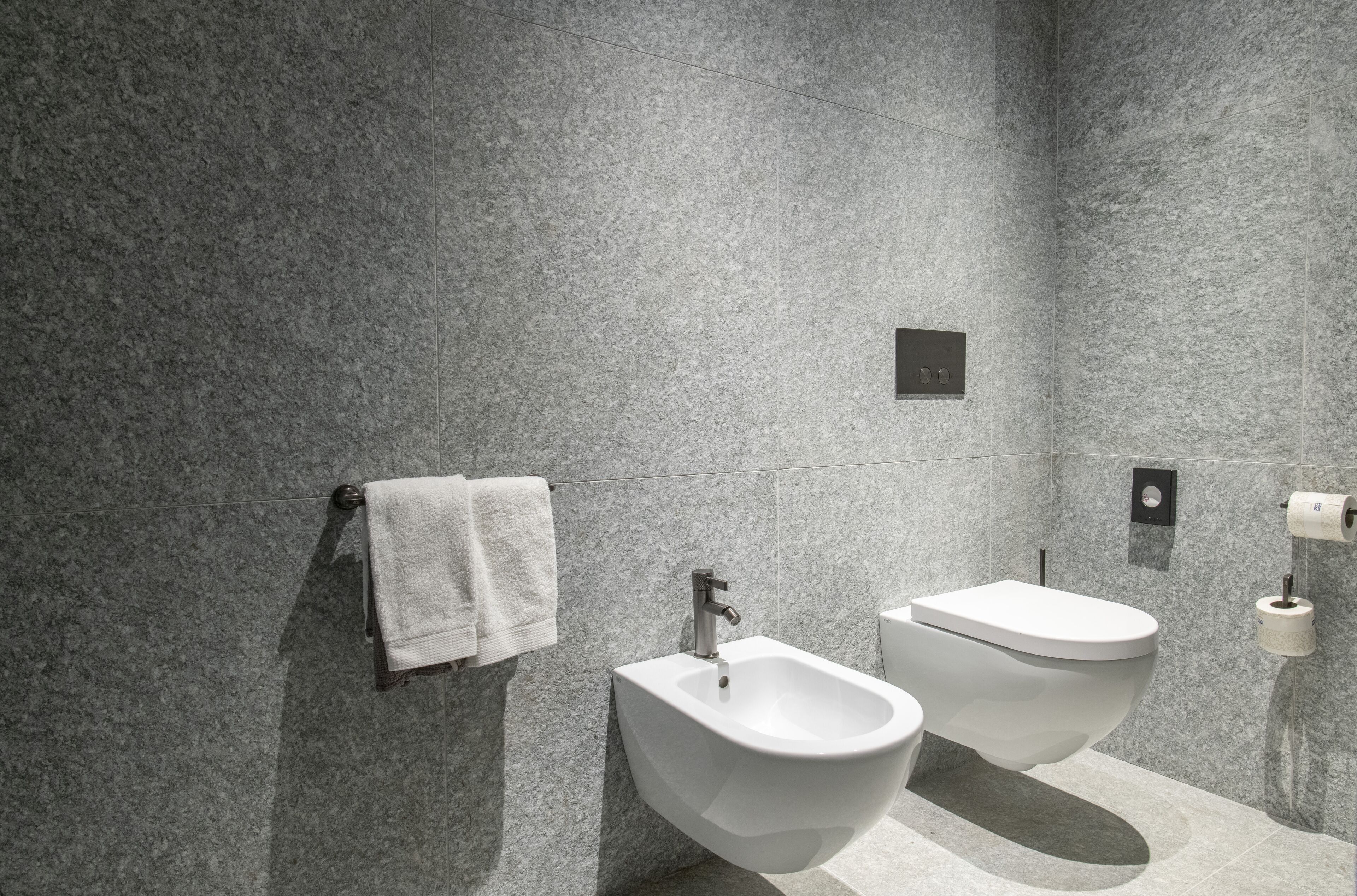 Et moderne badeværelse med bidét, toilet og håndklæderække foran en væg med struktur.
