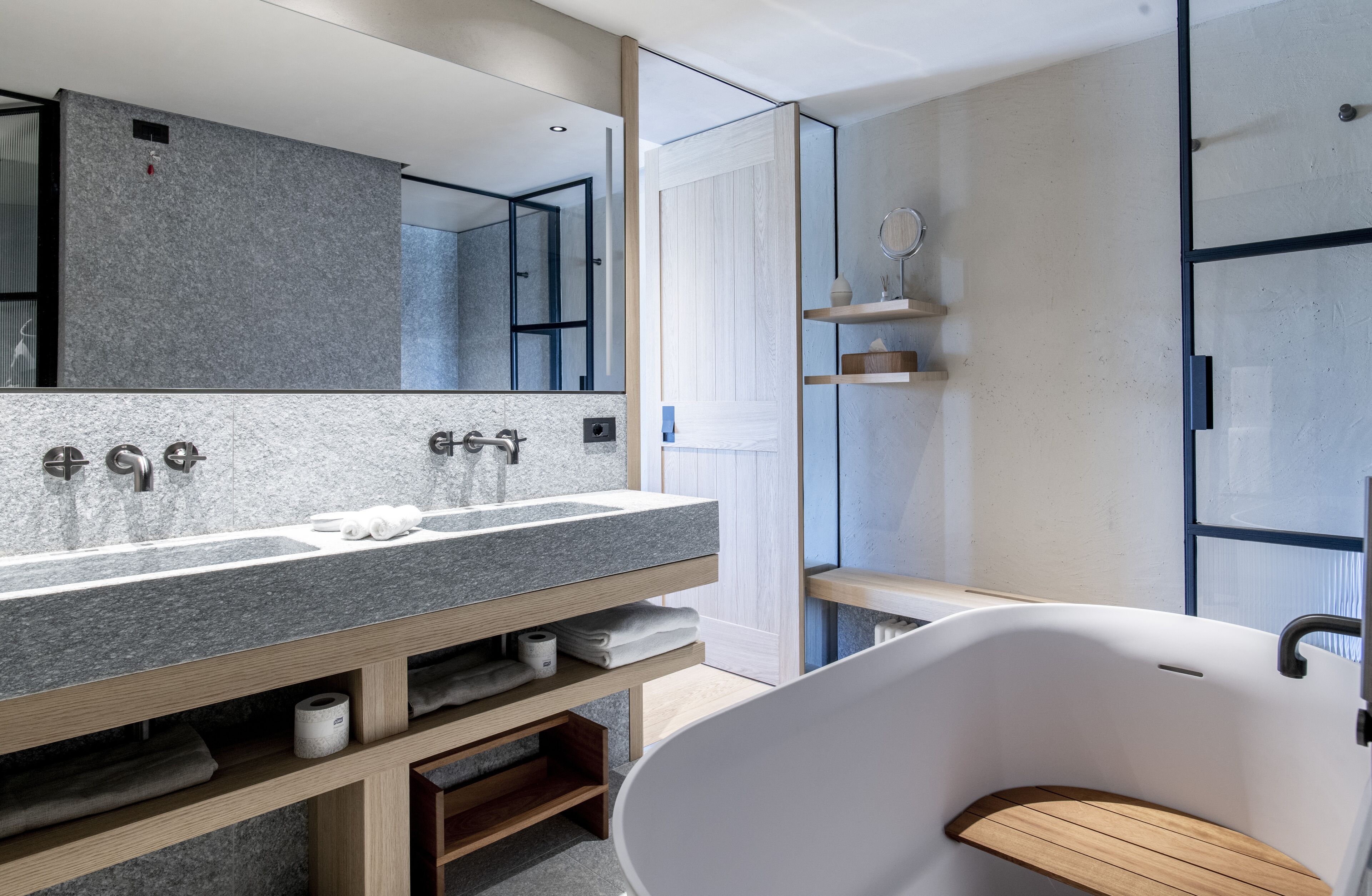 A modern bathroom with a granite countertop sink, a large mirror, a freestanding bathtub, and wooden shelves.