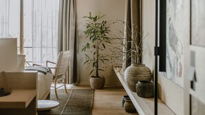 Una sala de estar moderna con techo alto, un ventanal con cortinas, un sofá, una silla, una planta en maceta y una estantería con objetos decorativos.