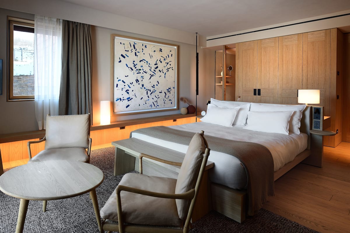 Une chambre d’hôtel avec un grand lit, deux fauteuils, une petite table, un grand tableau abstrait et une armoire en bois.