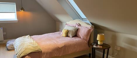 A bedroom with a sloped ceiling, a bed with pink bedding, a bedside lamp, and a window providing natural light.