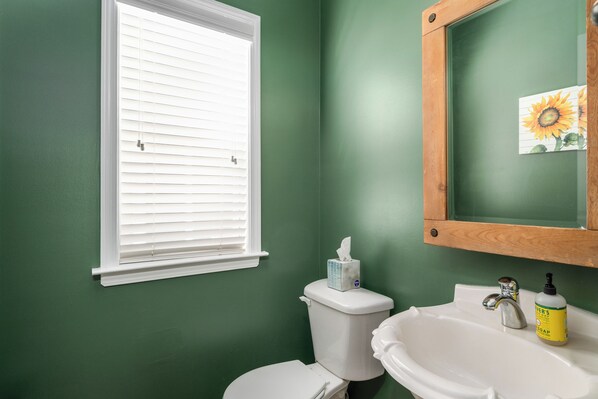 A bathroom with green walls, a white toilet, a sink with a mirror, and a window with blinds.
