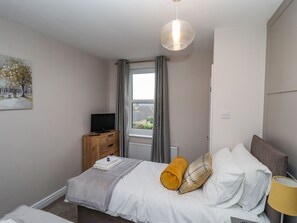 A bedroom with a bed, a TV on a wooden dresser, a window with curtains, and a painting on the wall. - Millside (Lincoln)