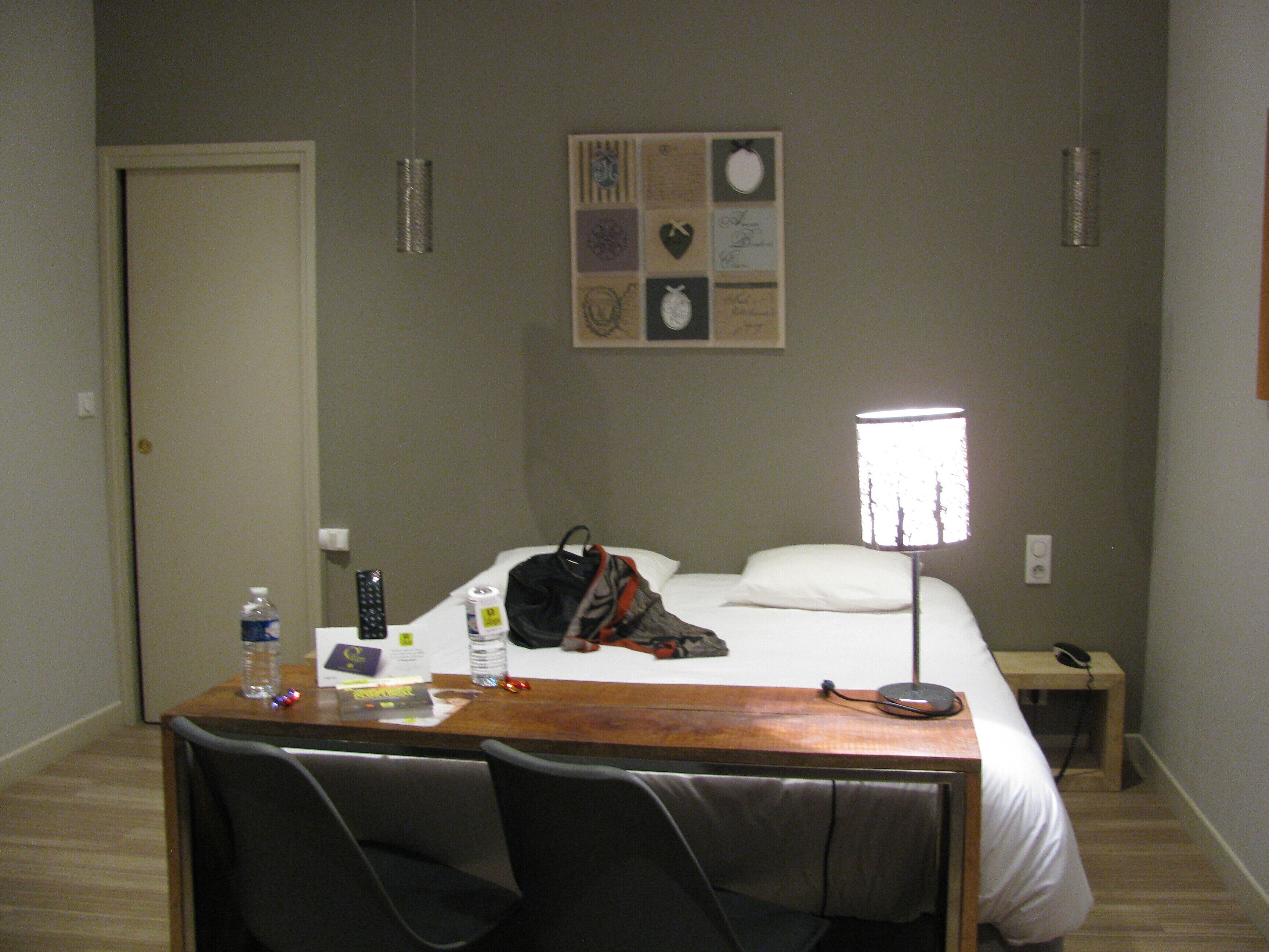 Een hotelkamer met een bed, een bureau met stoel, een nachtkastje en een wandklok.