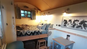 Ein gemütliches Zimmer mit einem Bett, einem kleinen Tisch mit Hocker, einem Sessel und einem an der Wand befestigten Regal.