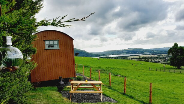 Eine Holzhütte mit kleinem Fenster, einem Picknicktisch und Blick auf ein grünes Feld und Hügel im Hintergrund.