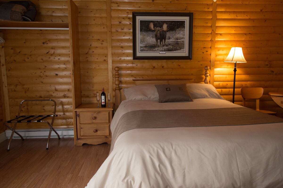 Een gezellig houten blokhut slaapkamer met een bed, een nachtkastje met een fles en glazen, een houten stoel en een afgebakend rendier beeld op de muur.
