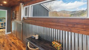 Ein rustikales Blockhaus-Interieur mit Holzwänden, einem großen Fenster mit Blick auf eine Berglandschaft, einer hölzernen Bank und einem kleinen Tisch mit Gegenständen darauf.