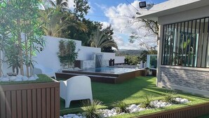 A modern outdoor patio with artificial grass, a small waterfall feature, and a seating area with white chairs. - Beautiful home 10 min from everythin!! (luquillo)