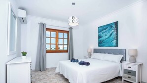 Ein Schlafzimmer mit einem Bett, Nachttischen, Vorhängen an einem Fenster sowie einer Wand-Klimaanlage und einem Bild.