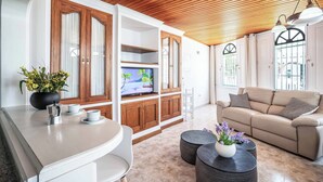 Ein modernes Wohnzimmer mit einem beigefarbenen Sofa, einem Fernseher, Holzschränken und einer Küchenzeile mit weißer Arbeitsplatte.